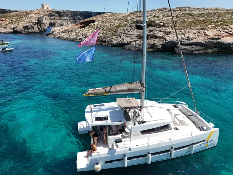 Angelina Mia catamaran at anchor in turquoise water below Comino's Santa Marija Tower